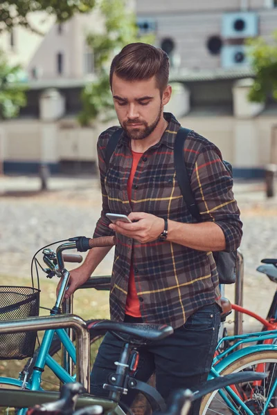 Un guapo hombre joven de moda con corte de pelo y barba con estilo, con camisa polar, se encuentra en el estacionamiento para bicicletas en el parque . —  Fotos de Stock