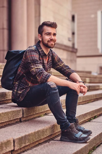 Hermoso hombre con corte de pelo elegante y barba, con una camisa de lana y vaqueros, sentado en los escalones contra un edificio viejo . —  Fotos de Stock