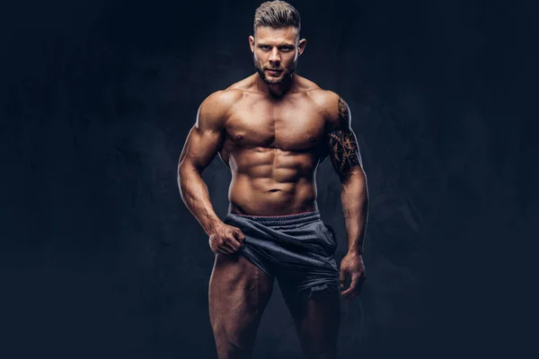 En stilig bar överkropp tatuerade bodybuilder med snygg frisyr och skägg, bära sport shorts, poserar i en studio. Isolerad på en mörk bakgrund — Stockfoto