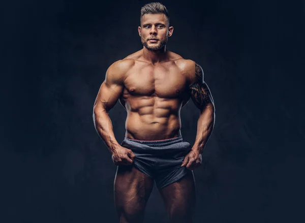A handsome shirtless tattooed bodybuilder with stylish haircut and beard, wearing sports shorts, posing in a studio. Isolated on a dark background — Stock Photo, Image