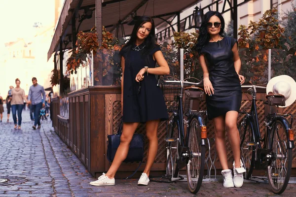 Twee sexy brunette dragen stijlvolle zwarte jurkjes, poseren in de buurt van fietsen tegen een achtergrond van een café. — Stockfoto