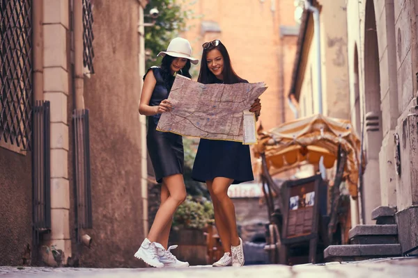 Dos mujeres turistas están de pie en medio de una calle estrecha, y buscan lugares de interés en el mapa . —  Fotos de Stock