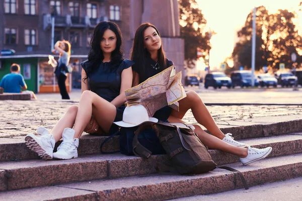 Duas mulheres turistas, relaxante depois de um passeio na mira, olhar em um mapa, sentado em degraus de pedra contra um fundo de pôr do sol brilhante . — Fotografia de Stock