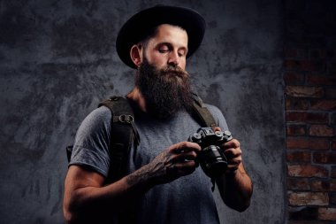 Portre yakışıklı bir sakallı gezgin bir sırt çantası ve dövmeli kolları ile şapkalı tutan karanlık bir arka plan üzerinde izole bir fotoğraf makinesi.