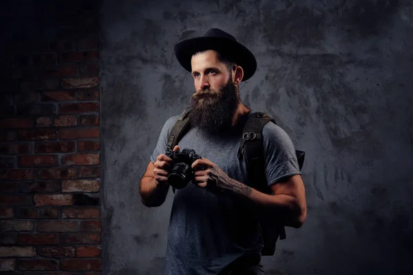 Retrato de un guapo viajero barbudo en un sombrero con una mochila y brazos tatuados, sostiene una cámara fotográfica, aislada sobre un fondo oscuro . — Foto de Stock