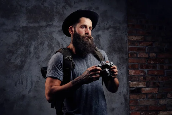 Retrato de un guapo viajero barbudo en un sombrero con una mochila y brazos tatuados, sostiene una cámara fotográfica, aislada sobre un fondo oscuro . — Foto de Stock