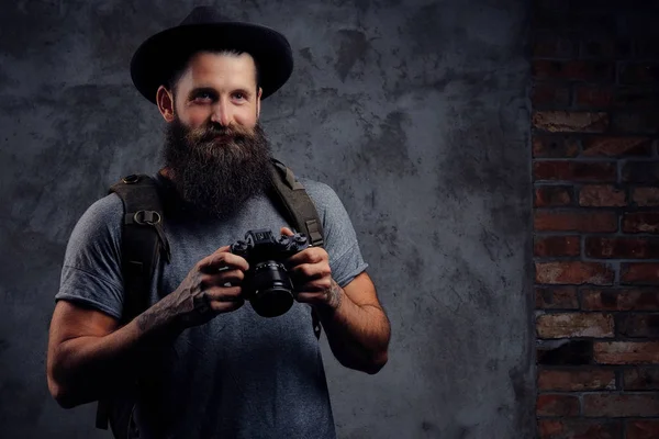 Retrato de um belo viajante barbudo em um chapéu com uma mochila e braços tatuados, segura uma câmera fotográfica, isolada em um fundo escuro . — Fotografia de Stock