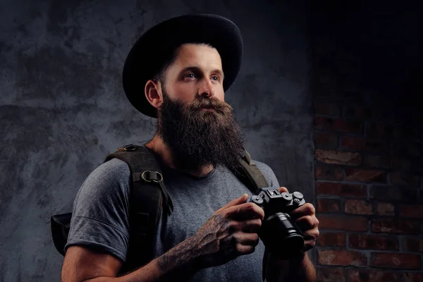 Retrato de un guapo viajero barbudo en un sombrero con una mochila y brazos tatuados, sostiene una cámara fotográfica, aislada sobre un fondo oscuro . —  Fotos de Stock