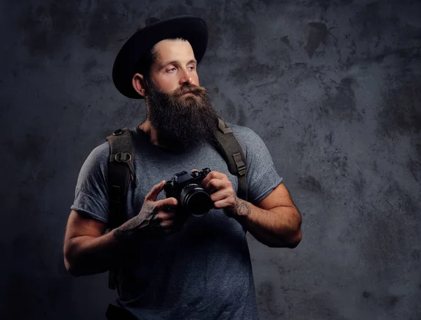 Retrato de un guapo viajero barbudo en un sombrero con una mochila y brazos tatuados, sostiene una cámara fotográfica, aislada sobre un fondo oscuro . — Foto de Stock