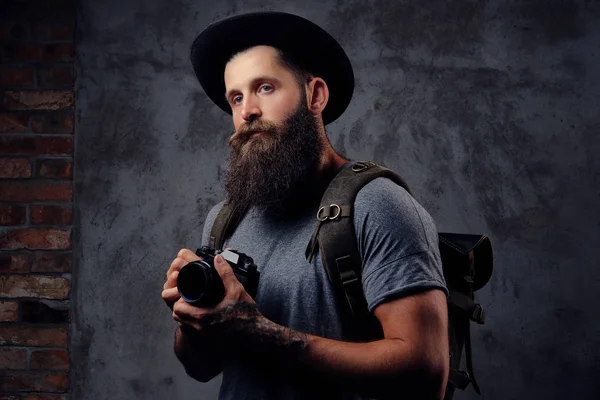 Retrato de um belo viajante barbudo em um chapéu com uma mochila e braços tatuados, segura uma câmera fotográfica, isolada em um fundo escuro . — Fotografia de Stock