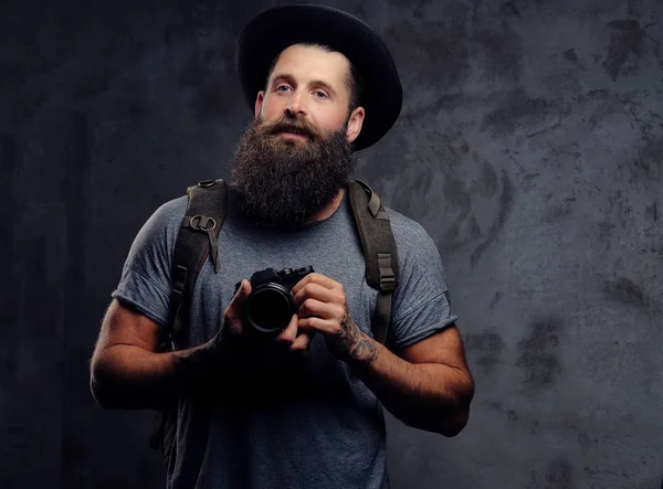 Retrato de un guapo viajero barbudo en un sombrero con una mochila y brazos tatuados, sostiene una cámara fotográfica, aislada sobre un fondo oscuro . — Foto de Stock