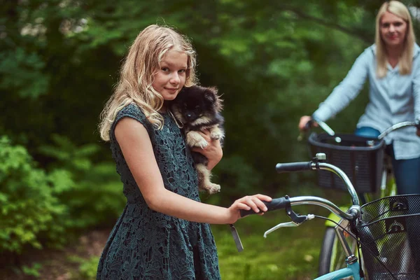 Porträtt av en mor och dotter med en blond hår på en cykel rida med sin söta lilla spitz hund i en park. — Stockfoto