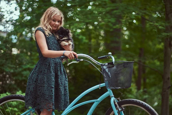 Retrato de una niña rubia en un vestido casual, sostiene lindo perro spitz, en un parque . — Foto de Stock