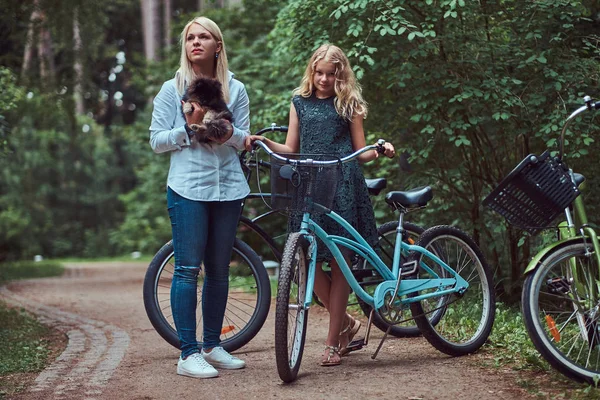 Porträt einer Mutter und einer Tochter mit blonden Haaren auf einer Fahrradtour mit ihrem niedlichen kleinen Spitzhund im Park. — Stockfoto