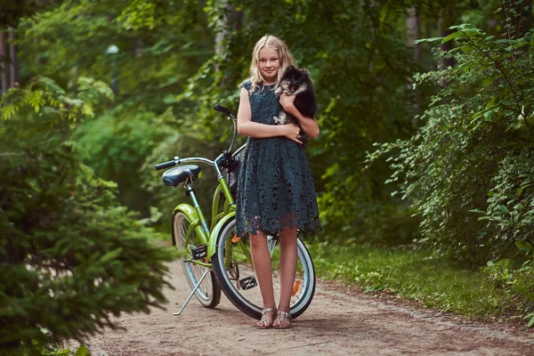 Ganzkörperporträt eines lächelnden kleinen blonden Mädchens in einem lässigen Kleid, hält niedlichen Spitz-Hund. Fahrradtour im Park. — Stockfoto