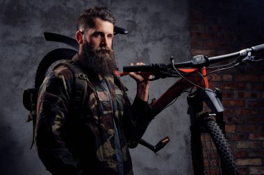 Yakışıklı bir gezgin, spor bisiklet bir stüdyoda bir omuz ile ayakta bir trendy Kamuflaj ceketi giymiş sakallı. Karanlık bir arka plan üzerinde izole.