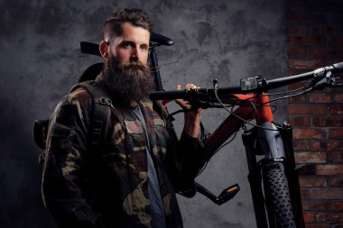 Yakışıklı bir gezgin, spor bisiklet bir stüdyoda bir omuz ile ayakta bir trendy Kamuflaj ceketi giymiş sakallı. Karanlık bir arka plan üzerinde izole.