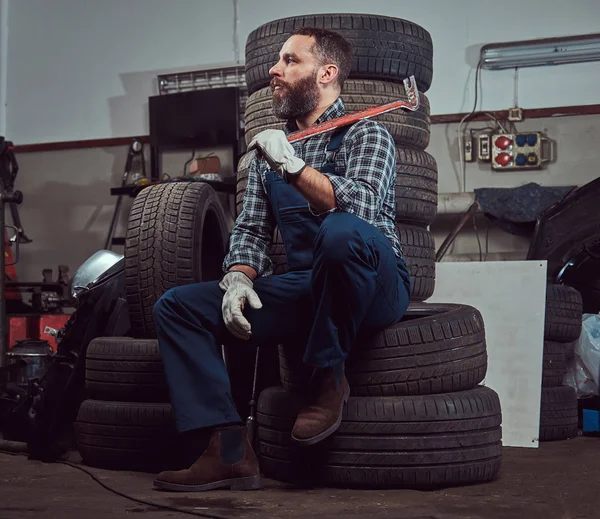 穿着制服的大胡子技工 拿着一把撬棍 坐在车库里的旧汽车轮胎上 — 图库照片