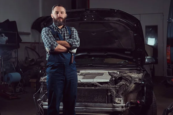 Bearded expert mechanic dressed in a uniform, standing with crossed arms against a car in the garage.