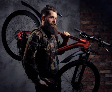 Yakışıklı bir gezgin, spor bisiklet bir stüdyoda bir omuz ile ayakta bir trendy Kamuflaj ceketi giymiş sakallı. Karanlık bir arka plan üzerinde izole.