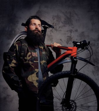 Yakışıklı bir gezgin, spor bisiklet bir stüdyoda bir omuz ile ayakta bir trendy Kamuflaj ceketi giymiş sakallı. Karanlık bir arka plan üzerinde izole.
