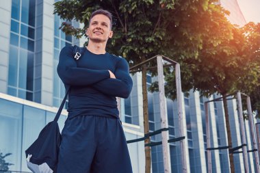 Modern kentin bir gökdelen karşı çapraz silah ile duran siyah bir spor giyim mutlu bir yakışıklı adam.