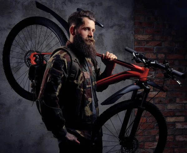 ハンサムなひげを生やした旅行者 スタジオで肩にスポーツ自転車の側に立って 流行の偽装ジャケットに身を包んだ 暗い背景に分離 — ストック写真