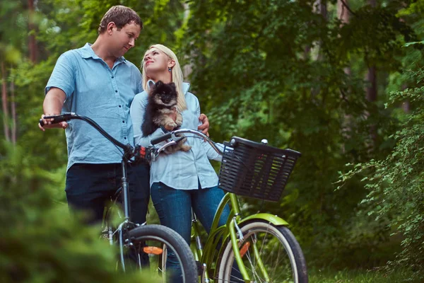 一个漂亮的夫妇的金发女郎和男子穿着休闲衣服骑自行车与他们可爱的小斯皮茨 — 图库照片