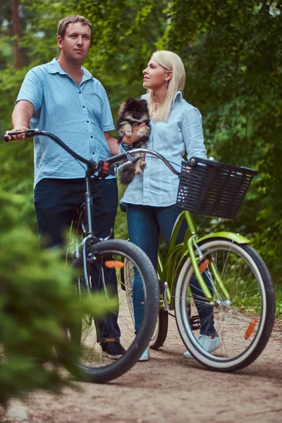 ブロンドの女性と男性の魅力的なカップルが かわいい小さなスピッツと自転車に乗ってカジュアルな服を着た — ストック写真