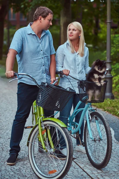 ブロンドの女性と男性の魅力的なカップル服を着たカジュアルな自転車に乗って かわいい小さなスピッツとかごの中 — ストック写真