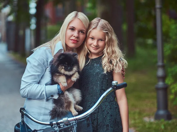 公園でかわいい小さなスピッツ犬と一緒に乗る母と自転車に乗って金髪娘の肖像 — ストック写真