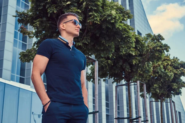 一个年轻英俊的时尚男士 戴着墨镜 穿着黑色 T恤和裤子 站在现代城市反对摩天大楼 — 图库照片