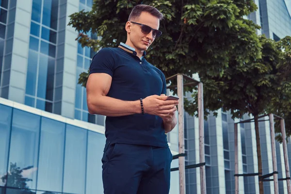 A handsome fashionable man with a stylish haircut in sunglasses, dressed in a black t-shirt and pants, reads a message on the smartphone, stans in the modern city against a skyscraper.