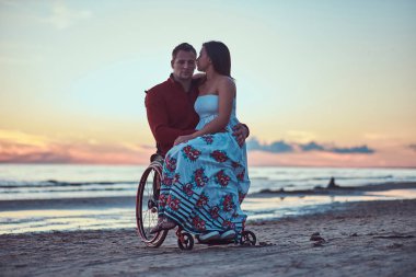 A loving couple, a woman sits on her husbands lap, resting on a beach against a background of a bright dawn. clipart