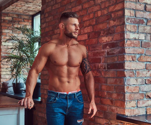Retrato de un hombre fuerte y guapo sin camisa con cuerpo musculoso, con jeans, apoyado en una pared de ladrillo . — Foto de Stock