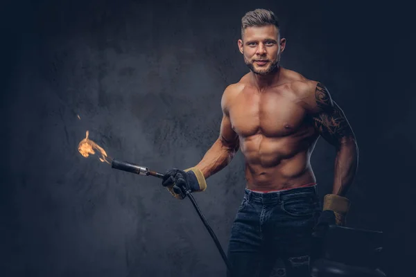 Brutal tattoed male welder with a stylish haircut and beard, with muscular body, dressed in only jeans, holds propane tank and a burning burner, standing in a studio, looking at a camera. — Stock Photo, Image