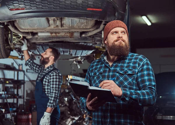 Twee bebaarde brute mechanica repareren een auto op een lift in de garage. — Stockfoto