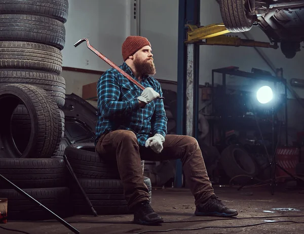 Un mecánico barbudo vestido con un uniforme, se sienta en neumáticos viejos del coche en el garaje . —  Fotos de Stock