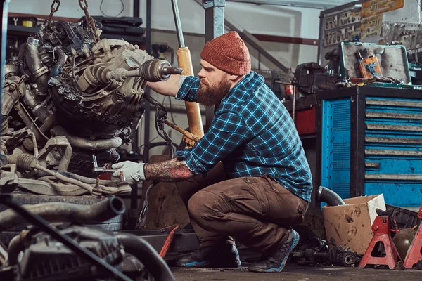 Un brutal especialista mecánico barbudo tatuado repara el motor del coche que se eleva en el ascensor hidráulico en el garaje. Estación de servicio . —  Fotos de Stock