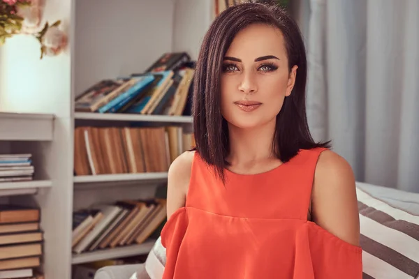 Retrato de uma bela morena feminina vestindo um vestido vermelho, sentada em um sofá . — Fotografia de Stock