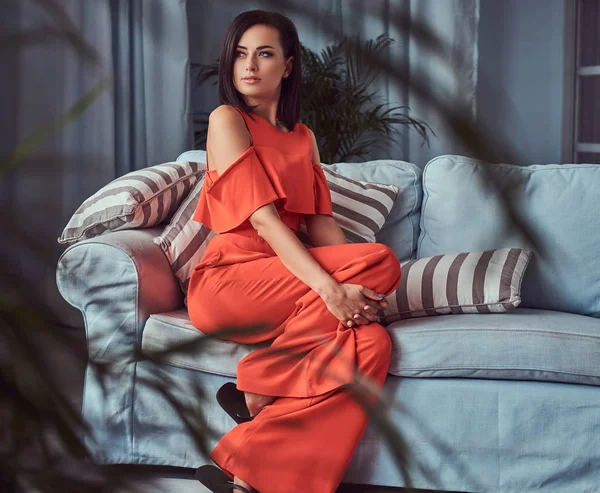 Retrato de uma bela morena feminina vestindo um vestido vermelho, sentada em um sofá . — Fotografia de Stock