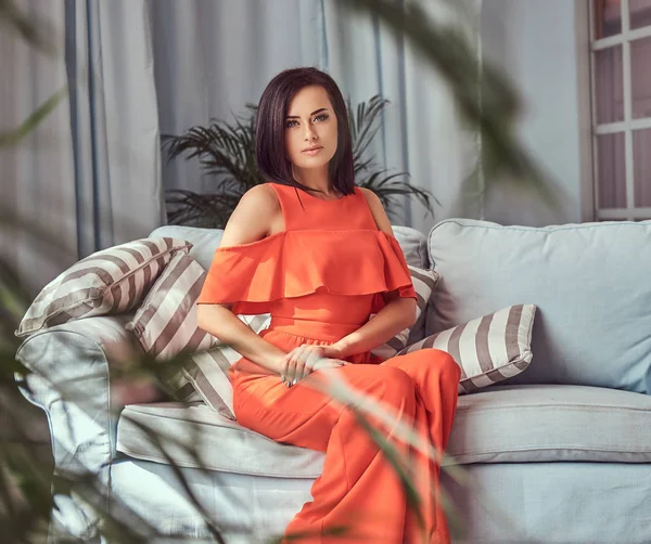 Retrato de uma bela morena feminina vestindo um vestido vermelho, sentada em um sofá . — Fotografia de Stock