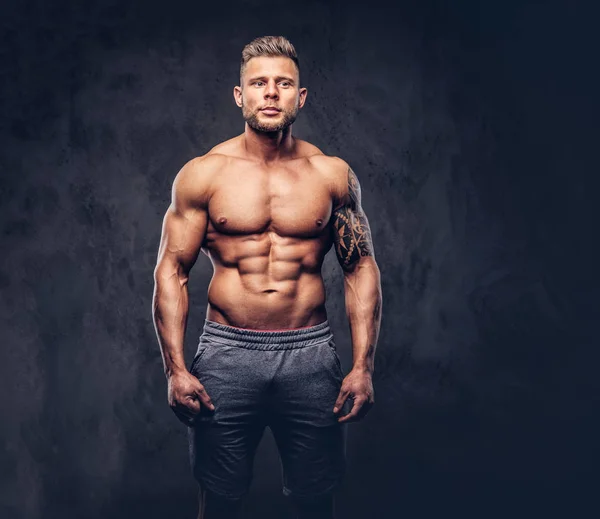 En stilig bar överkropp tatuerade bodybuilder med snygg frisyr och skägg, bära sport shorts, poserar i en studio. Isolerad på en mörk bakgrund — Stockfoto