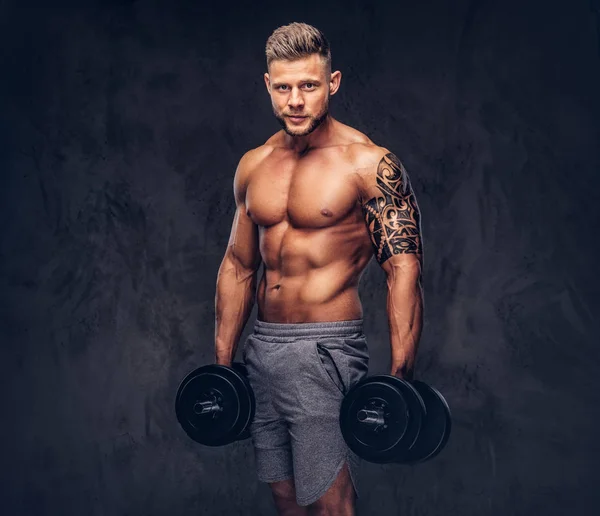 A handsome shirtless tattooed bodybuilder with stylish haircut and beard, wearing sports shorts, posing in a studio. Isolated on a dark background — Stock Photo, Image