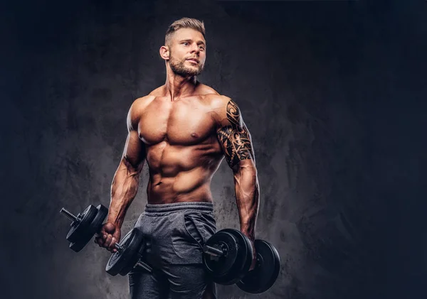 Un guapo culturista tatuado sin camisa con corte de pelo y barba con estilo, con pantalones cortos deportivos, posando en un estudio. Aislado sobre un fondo oscuro —  Fotos de Stock