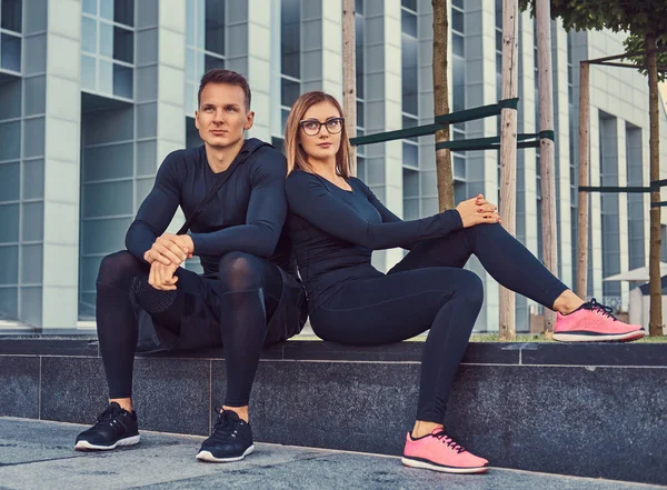 Fitness couple in a sportswear, sexy blonde girl and a handsome muscular guy sitting on a bench against a skyscraper. — Stock Photo, Image