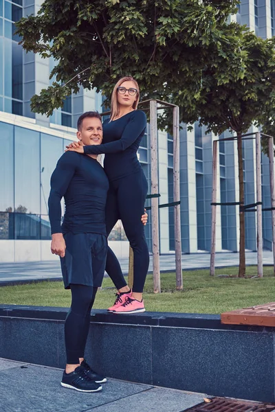 Happy fitness couple in a sportswear, sexy blonde girl and a handsome muscular guy stands in the modern city against a skyscraper. — Stock Photo, Image
