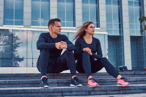 Um jovem par de aptidão em um sportswear, senta-se em passos na cidade moderna contra um arranha-céu. Conceito de estilo de vida saudável . — Fotografia de Stock