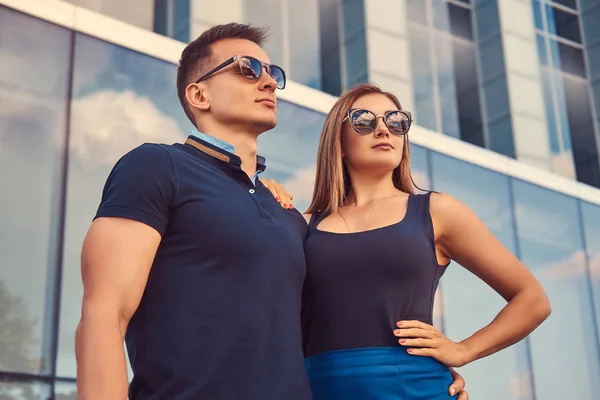 Attraktiva fashionabla par, sexig blond tjej och stilig man stående i den moderna staden mot en skyskrapa. — Stockfoto