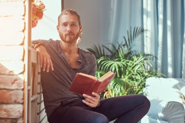 Yakışıklı şık Kızıl saçlı öğrenci içinde rahat giysiler, bir sandalye bir evde oturan ve bir kitap, bir kameraya bakıyor tutar.